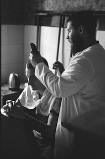 A black and white photograph capturing a barber giving a haircut to a seated client. The barber is focused, holding clippers close to the client's head. The client is sitting calmly with fingers interlaced. The setting appears to be inside a room with tiled walls, and a kettle is visible in the background.