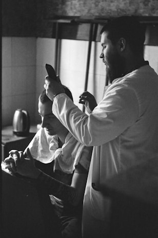 A black and white photograph capturing a barber giving a haircut to a seated client. The barber is focused, holding clippers close to the client's head. The client is sitting calmly with fingers interlaced. The setting appears to be inside a room with tiled walls, and a kettle is visible in the background.