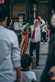 A street performer with a red nose is interacting with a puppet that has a green face and is wearing a trench coat and black hat. The performer is dressed in a white shirt and red tie. Several onlookers, including a child, are visible in the foreground, while a person in the background is wearing a mask. The scene appears to be taking place in an urban setting with shops and signs around.