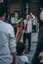 A street performer with a red nose is interacting with a puppet that has a green face and is wearing a trench coat and black hat. The performer is dressed in a white shirt and red tie. Several onlookers, including a child, are visible in the foreground, while a person in the background is wearing a mask. The scene appears to be taking place in an urban setting with shops and signs around.
