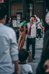 A street performer with a red nose is interacting with a puppet that has a green face and is wearing a trench coat and black hat. The performer is dressed in a white shirt and red tie. Several onlookers, including a child, are visible in the foreground, while a person in the background is wearing a mask. The scene appears to be taking place in an urban setting with shops and signs around.