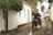 A cobblestone street lined with white colonial-style buildings, some with blue and yellow doors. A horse-drawn carriage is moving down the street, accompanied by people walking. Lush greenery hangs over the street from the rooftops and walls.
