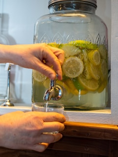A hand is pouring a beverage from a glass dispenser filled with sliced lemons and limes into a clear glass. The dispenser has a silver spout and is positioned on a wooden surface.