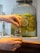 A hand is pouring a beverage from a glass dispenser filled with sliced lemons and limes into a clear glass. The dispenser has a silver spout and is positioned on a wooden surface.