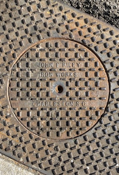 A close-up view of a metal manhole cover with a grid pattern. The cover has the text 'JOHN B. RILEY IRON WORKS CHARLESTON S.C.' embossed on it. The surrounding surface also has a textured grid pattern and appears weathered.
