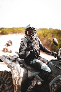 A person is wearing protective gear, including a helmet and visor, riding an all-terrain vehicle (ATV) on a sandy terrain. The ATV and the rider's clothing are dusty, suggesting off-road adventure. The background features green shrubs under a bright sky.