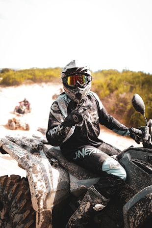 A person is wearing protective gear, including a helmet and visor, riding an all-terrain vehicle (ATV) on a sandy terrain. The ATV and the rider's clothing are dusty, suggesting off-road adventure. The background features green shrubs under a bright sky.