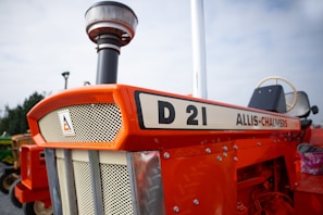 A vintage Allis-Chalmers D21 tractor with a bright orange exterior is prominently displayed. The front grille features an emblem, and there is visible detailing with bolts and metallic finishes. The surrounding environment is outdoors, with a clear sky in the background and other agricultural equipment visible in the distance.