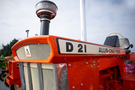 A vintage Allis-Chalmers D21 tractor with a bright orange exterior is prominently displayed. The front grille features an emblem, and there is visible detailing with bolts and metallic finishes. The surrounding environment is outdoors, with a clear sky in the background and other agricultural equipment visible in the distance.