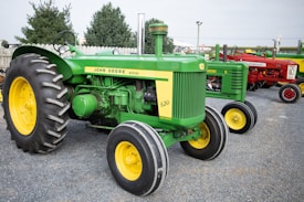 A row of vintage tractors is displayed on a gravel lot, featuring prominently a green and yellow John Deere diesel tractor with the model number 820. In the background, other red and green tractors are visible, along with trees and a wooden fence.