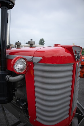 A close-up of a vintage tractor, highlighting its bright red paint and metal grille front. The design features a distinctive round headlight on the side and a chrome exhaust pipe. The tractor's badge is prominently displayed on the front, and the background is slightly blurred, indicating an outdoor setting.