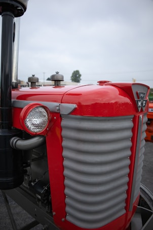 A close-up of a vintage tractor, highlighting its bright red paint and metal grille front. The design features a distinctive round headlight on the side and a chrome exhaust pipe. The tractor's badge is prominently displayed on the front, and the background is slightly blurred, indicating an outdoor setting.