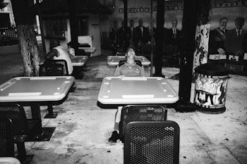 A person is sitting at a table in an outdoor area. The scene includes multiple empty tables and chairs, with one person casually seated in a hat and casual attire. In the background, there is a mural featuring images of several people. A trash can with the slogan 'Keep Miami Beach Clean' is positioned to the right.