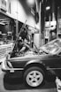A mechanic is working on a car in a garage. The hood of the car is open, and the mechanic is reaching into the engine compartment. The environment is industrial, with various tools and equipment visible in the background. The scene is captured in black and white, providing a classic and timeless feel.