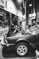 A mechanic is working on a car in a garage. The hood of the car is open, and the mechanic is reaching into the engine compartment. The environment is industrial, with various tools and equipment visible in the background. The scene is captured in black and white, providing a classic and timeless feel.
