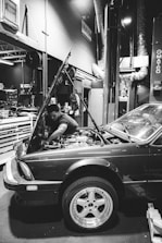 A mechanic is working on a car in a garage. The hood of the car is open, and the mechanic is reaching into the engine compartment. The environment is industrial, with various tools and equipment visible in the background. The scene is captured in black and white, providing a classic and timeless feel.
