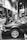 A mechanic is working on a car in a garage. The hood of the car is open, and the mechanic is reaching into the engine compartment. The environment is industrial, with various tools and equipment visible in the background. The scene is captured in black and white, providing a classic and timeless feel.