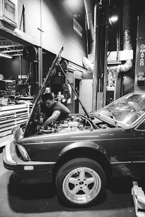 A mechanic is working on a car in a garage. The hood of the car is open, and the mechanic is reaching into the engine compartment. The environment is industrial, with various tools and equipment visible in the background. The scene is captured in black and white, providing a classic and timeless feel.