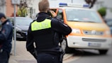 A person in a police uniform is speaking on a phone, facing away from the camera. A police vehicle with an orange stripe is parked nearby.