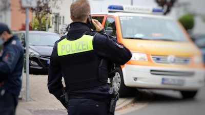 A person in a police uniform is speaking on a phone, facing away from the camera. A police vehicle with an orange stripe is parked nearby.