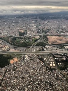 Aerial view of a sprawling urban landscape, featuring densely packed buildings, highways, and patches of greenery. The city is illuminated by a soft, diffused light under a cloudy sky, with visible infrastructure such as roads and industrial areas.