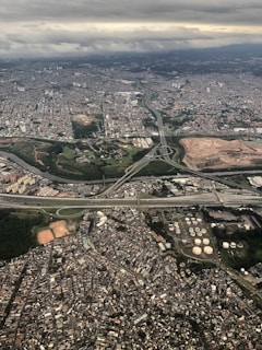 Aerial view of a sprawling urban landscape, featuring densely packed buildings, highways, and patches of greenery. The city is illuminated by a soft, diffused light under a cloudy sky, with visible infrastructure such as roads and industrial areas.