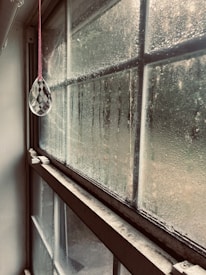 A window is covered in condensation, with droplets trailing down the glass panes. A clear, faceted glass ornament hangs from a pink string, catching the light softly. The window frame appears old and weathered, with visible dust and grime along the edges. Outside, a blurred view suggests foliage or an outdoor scene.
