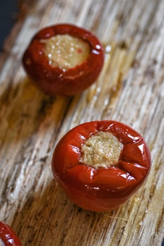 Two red round vegetables with a stuffing inside are placed on a rustic wooden surface. The vegetables' skins are slightly glossy, indicating that they may have been roasted or cooked. The filling appears to be a smooth, brownish mixture.