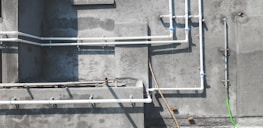 A concrete wall features several white PVC pipes running vertically and horizontally across its surface. Some pipes are connected by blue coupling, and a single bright green pipe or cable contrasts against the muted background. Shadows of the pipes are cast clearly on the wall, indicating sunlight. The textures of the concrete are visible, with some areas appearing rougher or stained.