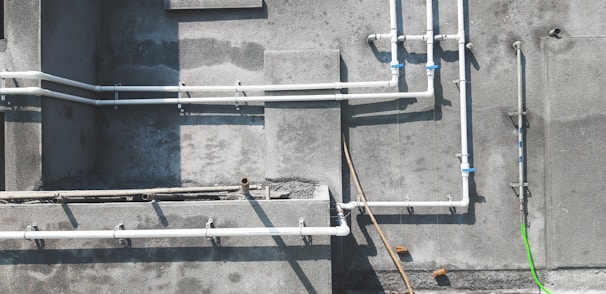 A concrete wall features several white PVC pipes running vertically and horizontally across its surface. Some pipes are connected by blue coupling, and a single bright green pipe or cable contrasts against the muted background. Shadows of the pipes are cast clearly on the wall, indicating sunlight. The textures of the concrete are visible, with some areas appearing rougher or stained.