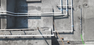 A concrete wall features several white PVC pipes running vertically and horizontally across its surface. Some pipes are connected by blue coupling, and a single bright green pipe or cable contrasts against the muted background. Shadows of the pipes are cast clearly on the wall, indicating sunlight. The textures of the concrete are visible, with some areas appearing rougher or stained.