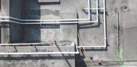 A concrete wall features several white PVC pipes running vertically and horizontally across its surface. Some pipes are connected by blue coupling, and a single bright green pipe or cable contrasts against the muted background. Shadows of the pipes are cast clearly on the wall, indicating sunlight. The textures of the concrete are visible, with some areas appearing rougher or stained.