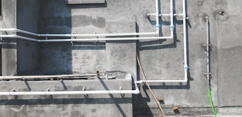 A concrete wall features several white PVC pipes running vertically and horizontally across its surface. Some pipes are connected by blue coupling, and a single bright green pipe or cable contrasts against the muted background. Shadows of the pipes are cast clearly on the wall, indicating sunlight. The textures of the concrete are visible, with some areas appearing rougher or stained.