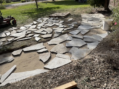 A partially constructed stone pathway laid out on a bed of sand and dirt with uneven edges. Large gray stones are scattered around, some already positioned while others are yet to be placed. In the background, there is a lawn mower on green grass, a wooden pallet, and a white bucket on the right side. The area appears to be a garden or yard under construction.