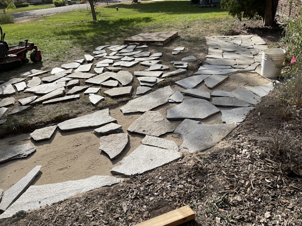 A partially constructed stone pathway laid out on a bed of sand and dirt with uneven edges. Large gray stones are scattered around, some already positioned while others are yet to be placed. In the background, there is a lawn mower on green grass, a wooden pallet, and a white bucket on the right side. The area appears to be a garden or yard under construction.