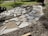 A partially constructed stone pathway laid out on a bed of sand and dirt with uneven edges. Large gray stones are scattered around, some already positioned while others are yet to be placed. In the background, there is a lawn mower on green grass, a wooden pallet, and a white bucket on the right side. The area appears to be a garden or yard under construction.