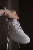 A hand with tattoos is holding a white sneaker with laces. The shoe features detailed stitching and a signature logo on the side. The background is a neutral dark shade, emphasizing the shoe's design.