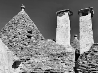 Conical stone roofs and cylindrical chimneys made from stacked stones characterize a traditional architectural style. The surface texture of the stones is rough, enhancing the rustic appearance. The monochromatic tone gives it a historical and timeless feel.