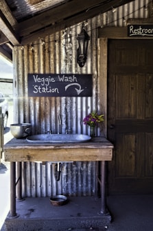 A rustic wooden sink station is mounted on a corrugated metal wall. Above it, a chalkboard sign reads 'Veggie Wash Station' with an arrow pointing downward. A metal strainer sits on the left side of the sink, while a small bouquet of colorful flowers is on the right. There is a dark wooden door with a 'Restroom' sign to the right, and an old-fashioned lantern is fixed to the wall above.