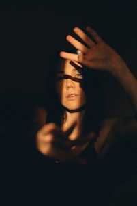 A person is partially obscured by shadows, with light casting across their face and hands. They appear to be reaching out with both hands, creating a dramatic play of light and darkness. The atmosphere is introspective and moody, with a strong contrast between illuminated and shadowed areas.