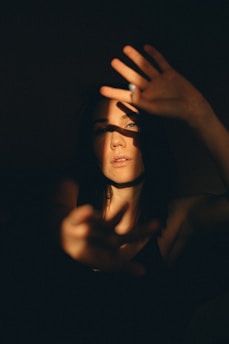 A person is partially obscured by shadows, with light casting across their face and hands. They appear to be reaching out with both hands, creating a dramatic play of light and darkness. The atmosphere is introspective and moody, with a strong contrast between illuminated and shadowed areas.