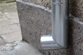 A metal downspout attached to the exterior of a building, directed at the pavement below. The pipe has a shiny, metallic finish and curves near the bottom.
