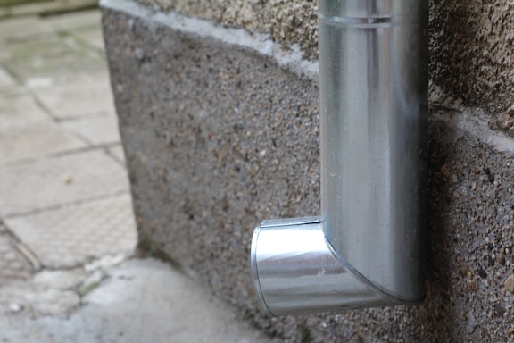 A metal downspout attached to the exterior of a building, directed at the pavement below. The pipe has a shiny, metallic finish and curves near the bottom.