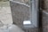 A metal downspout attached to the exterior of a building, directed at the pavement below. The pipe has a shiny, metallic finish and curves near the bottom.