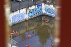 A construction site with graffiti-covered concrete walls and a partially flooded area. Several traffic cones are scattered in the water. The surrounding area suggests an industrial or urban setting, with debris and construction materials visible.