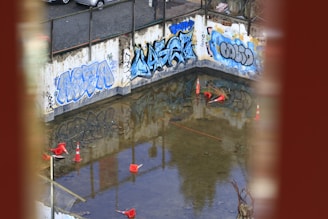 A construction site with graffiti-covered concrete walls and a partially flooded area. Several traffic cones are scattered in the water. The surrounding area suggests an industrial or urban setting, with debris and construction materials visible.