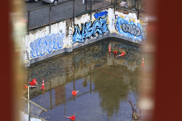 A construction site with graffiti-covered concrete walls and a partially flooded area. Several traffic cones are scattered in the water. The surrounding area suggests an industrial or urban setting, with debris and construction materials visible.