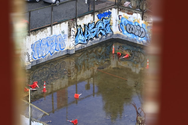 A construction site with graffiti-covered concrete walls and a partially flooded area. Several traffic cones are scattered in the water. The surrounding area suggests an industrial or urban setting, with debris and construction materials visible.