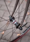 Close-up of a bicycle wheel, focusing on the hub and spokes with part of the tire visible. The pavement beneath features faint outlines of bricks. A few scattered brown leaves add a touch of autumn to the scene.