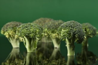 Several broccoli heads are standing upright on a reflective surface, creating a mirrored effect. The background is a solid, vibrant green, enhancing the fresh appearance of the broccoli.